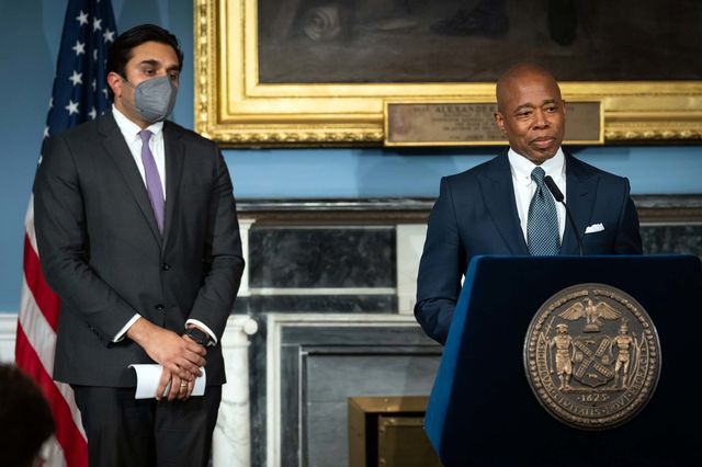 Health Commissioner Ashwin Vasan wears a mask on the left while Mayor Eric Adams, unmasked, speaks at a lectern in City Hall